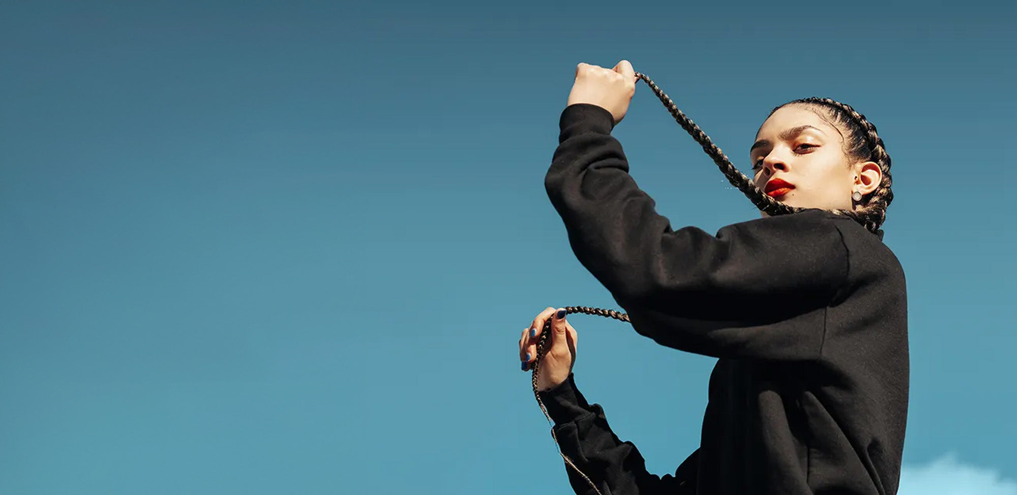 A woman turned to the side and holding her long black braids in front of a blue sky.