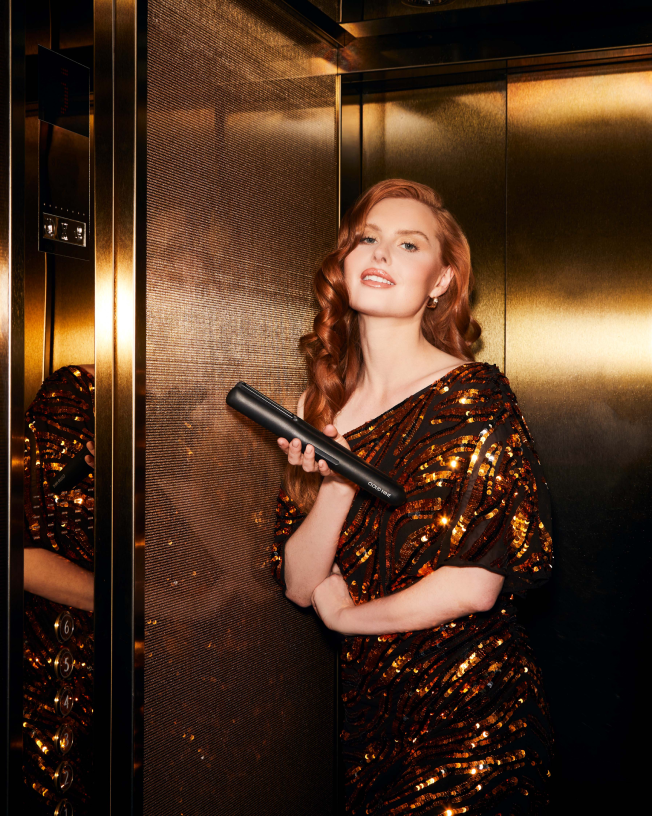 A smiling woman with red hair wearing an orange sequin dress, holding a CLOUD NINE straightener with a gold background. 